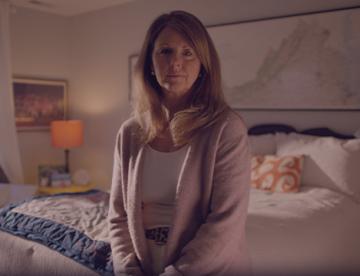 An image of Kerri Rhodes standing in a bedroom looking at the camera with a serious look